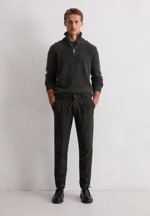 Donkergrijze ritsvest, groene broek en zwarte schoenen. Het vest heeft geribbelde mouwen en zoom, de broek heeft een rechte pasvorm en een knoopsluiting.