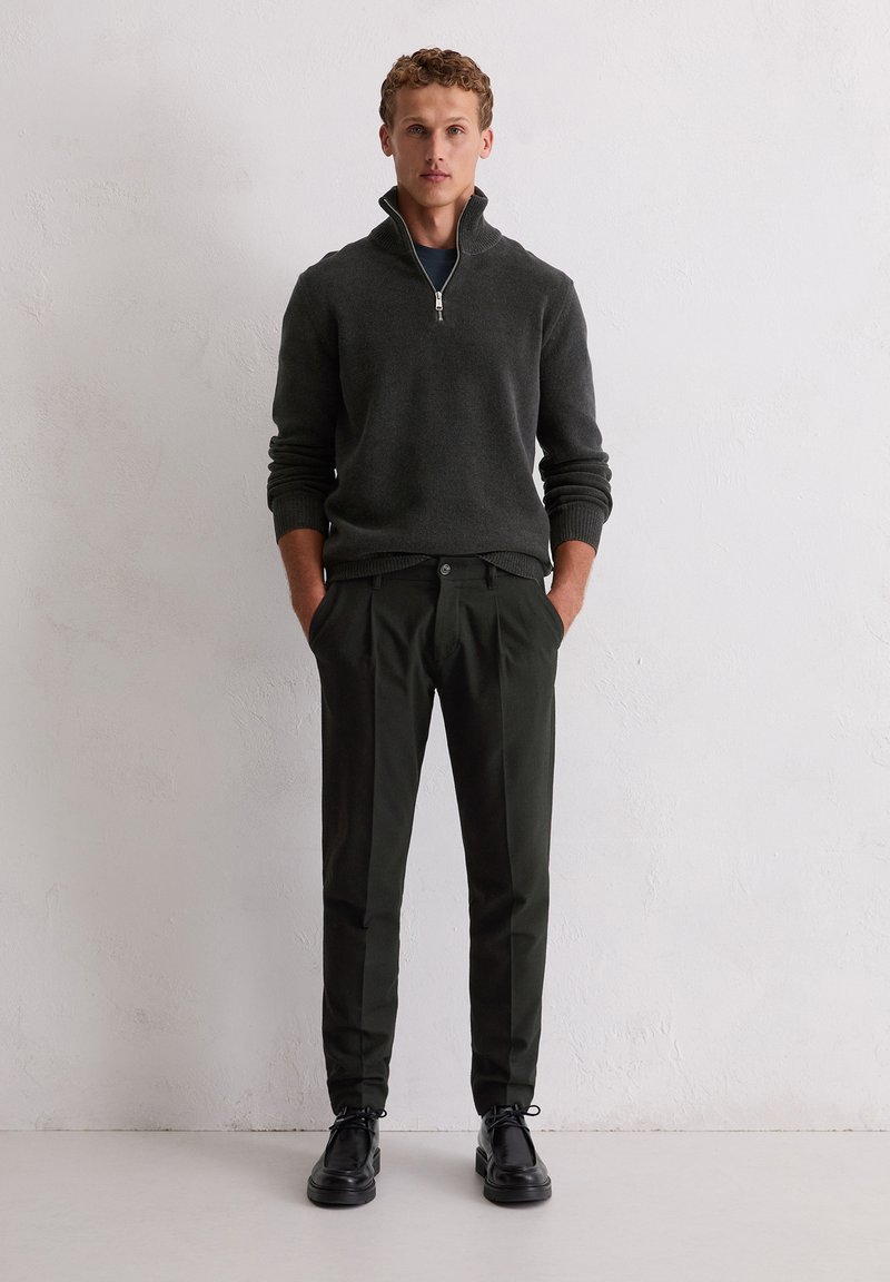 Donkergrijze ritsvest, groene broek en zwarte schoenen. Het vest heeft geribbelde mouwen en zoom, de broek heeft een rechte pasvorm en een knoopsluiting.
