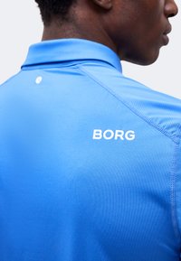 Άνδρας που φορά φωτεινό μπλε πόλο με το «BORG» τυπωμένο λευκό στο πάνω μέρος της πλάτης, πάνω σε απλό φωτεινό φόντο.