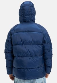 Osoba v modrej puffovej bundě s kapucí, pohled zezadu, v kombinaci se světle modrými džíny.