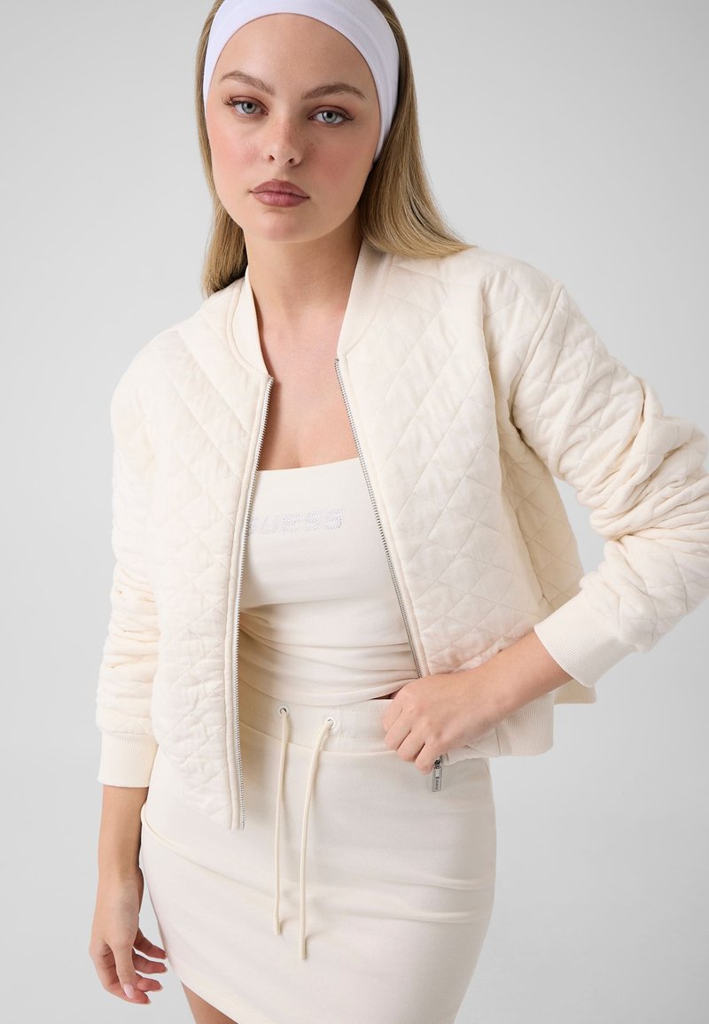 Jeune femme portant une veste matelassée blanche, une jupe assortie, un débardeur et un bandeau blanc sur un fond clair uni.