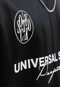Czarna tkanina z białym napisem „UNIVERSAL” oraz okrągłym logo z pionowymi stylizowanymi literami, obok srebrnego łańcuszka.