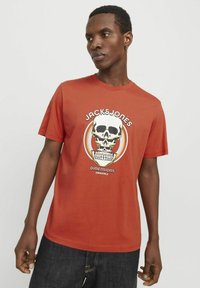 Crvena pamučna majica s grafičkim dizajnom lobanje oko koje se nalaze natpisi "JACK & JONES" i "DIMENSIONS". Kratki rukavi, okrugli ovratnik.