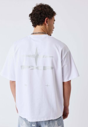Uomo con capelli ricci che indossa una T-shirt bianca oversize con una stella d'argento e la scritta "PXLP" sulla schiena, abbinata a jeans di denim chiaro.