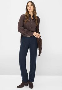 Donkerbruine satijnen button-up shirt, marineblauwe op maat gemaakte broek en bruine suède enkellaarzen. Model houdt een bruine franjes schoudertas vast.