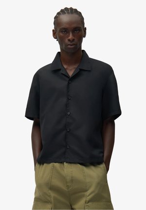 Giovane con capelli intrecciati che indossa una camicia nera a maniche corte con bottoni e pantaloni cargo beige, mani nelle tasche, espressione neutra.