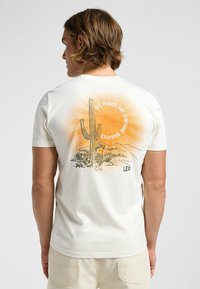 Camiseta de algodón blanca con un gráfico de un cactus y un niño contra un fondo de rayos solares en naranja y amarillo, con el texto "LEE HACE QUE EL SOL BRILLE MÁS".