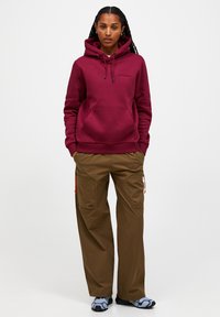Felpa maroon con cappuccio e cordini, con tasca frontale, abbinata a pantaloni cargo marroni con tasche con zip e dettagli arancioni.