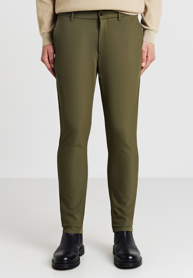 Olivgrüne Hosen mit schlanker Passform, aus einem glatten Stoff gefertigt. Verfügt über Standardtaschen und einen flachen Vorderteil, kombiniert mit schwarzen Knöchelstiefeln.