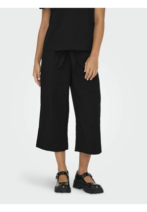 Femme portant un pantalon large noir court avec une taille nouée et des chaussures Mary Jane noires épaisses sur un fond uni.