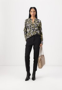 Svart og gul blomstrete bluse med lange ermer, brukt med skreddersydde svarte bukser og svarte ankelstøvler, holder en beige håndveske.