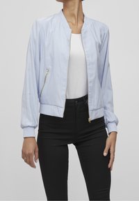 Světle modrá bomber bunda s předním zipem, pružnými manžetami a boční kapsou se zlatými komponenty. Spárována s černými džínami s vysokým pasem.