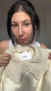 Mladá žena s tmavými vlasy drží krémový pletený oděv s ozdobným límcem a štítkem „YAS.“