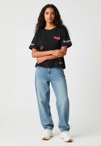 Melnas kokvilnas T-krekls ar grafikas drukām sarkanā, dzeltenā un baltā krāsā, apvienots ar gaiši ziliem platām biksēm un baltām tenisa kedām. Viens tonis fons.