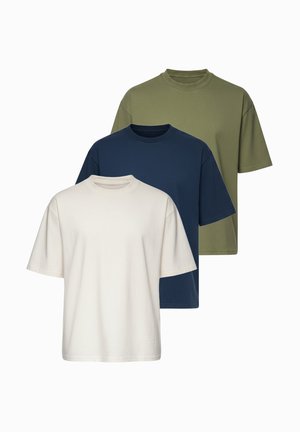 Man staat en draagt een loszittend beige t-shirt, lichtblauwe spijkerbroek, witte sneakers en een zilveren horloge op een witte achtergrond.