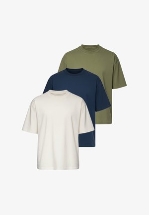 Man staat en draagt een loszittend beige t-shirt, lichtblauwe spijkerbroek, witte sneakers en een zilveren horloge op een witte achtergrond.