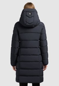 Palton lung, de tip puf, de culoare închisă, cu un model matlasat în formă de zigzag, cu glugă mare și detalii pe spate. Are un accent cu nasture în apropierea gâtului.