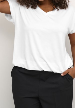 Persoon met krullend haar die een simpel wit T-shirt met korte mouwen draagt, ingestopt in een zwarte broek, met één hand in de zak en de arm omhoog.