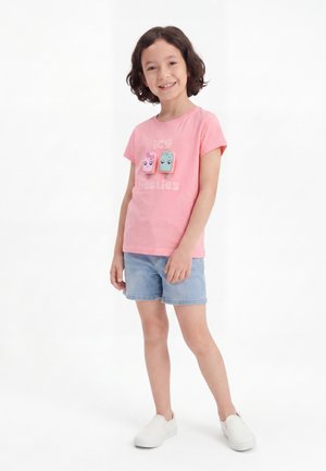 Smilende barn iført en lyserød T-shirt med tegneserie-ispinde og teksten "icy besties", denimshorts og hvide slip-on sko, stående på hvid baggrund.