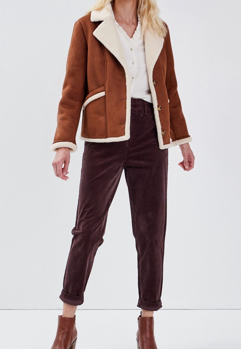 Femme portant une veste en peau retournée marron, un chemisier blanc boutonné, un pantalon en velours côtelé foncé avec les revers relevés, et des bottines marron, debout devant un fond uni.