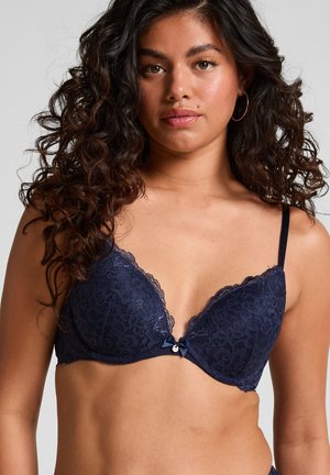 Vrouw met krullend, donker haar die een marineblauwe kanten beha draagt met dunne bandjes en een klein strikje in het midden aan de voorkant.