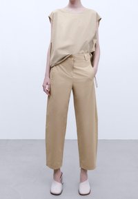 Top corto beige con vestibilità ampia, abbinato a pantaloni beige a gamba larga, dotati di chiusura a bottone e tasche laterali. Tessuto liscio e leggero.