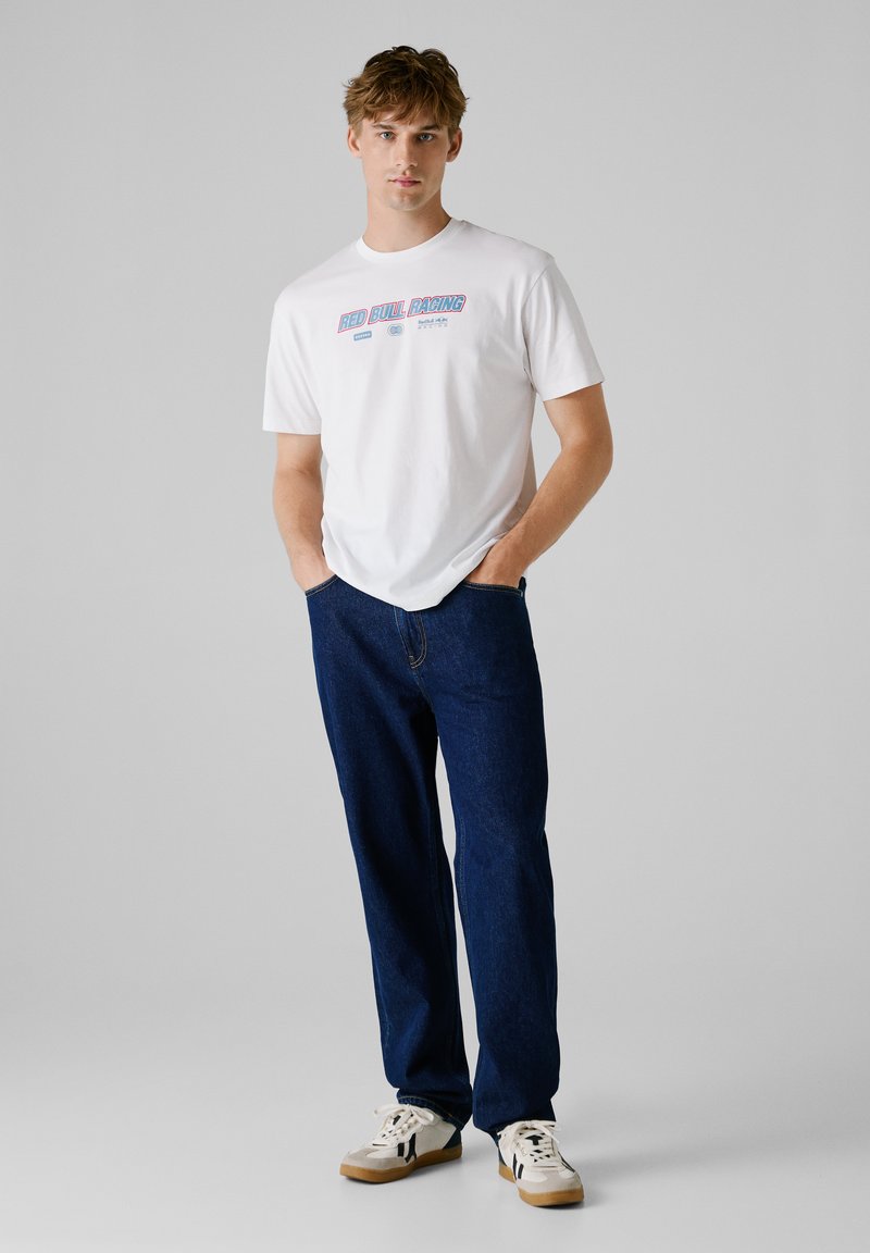 Biała bawełniana koszulka z grafiką "RED BULL RACING", zestawiona z ciemnoniebieskimi dżinsami i kremowymi sneakersami z czarnymi akcentami.