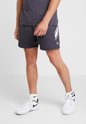 Mężczyzna ubrany w ciemne sportowe spodenki z białymi paskami, białe trampki Nike i białe skarpetki, stoi na jednolitym tle.