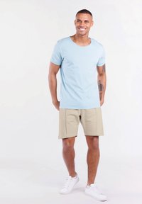 Hellblaue Baumwoll-T-Shirt mit rundem Halsausschnitt, kombiniert mit beigen Shorts mit genähten Akzenten. Weiße Sneakers vervollständigen das Outfit.