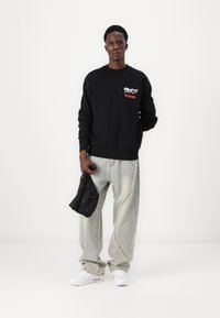 Sort sweatshirt med røde og hvide logoer, lysegrå slidte jeans med synlige sømme, og hvide sneakers. Holder en sort taske.