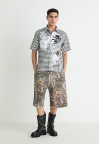 Košulja na kratke rukave s tartan uzorkom i crnom grafikom lubanje, u kombinaciji s kratkim hlačama u kamuflažnom uzorku i crnim čizmama do koljena s detaljima kopči.