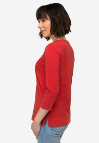 Camicia rossa a maniche lunghe con scollo rotondo, fessure laterali all'orlo, realizzata in morbido cotone. Indossata con jeans azzurri chiari.