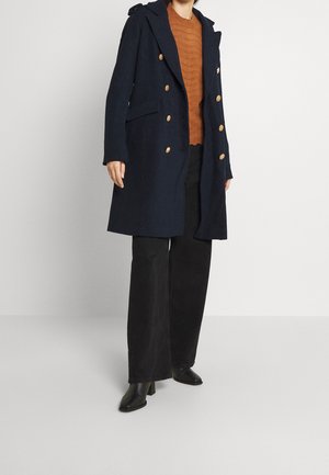 Marineblå dobbeltbrystet frakke med gyldne knapper, parret med en struktureret rustfarvet sweater og sorte vidde bukser samt højhælede sko.