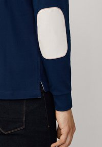 Marinefarbene Langarmshirt mit weißen Ellbogenpatches, kontrastierenden roten Nähten und seitlichen Schlitzen am Saum. Weiches, strukturiertes Material.