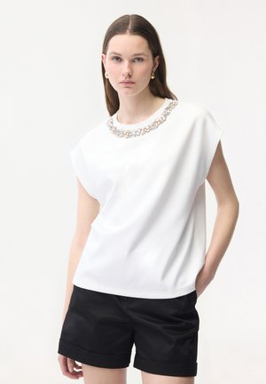 Młoda kobieta o brązowych włosach nosi białą, bezrękawnikową bluzkę ozdobioną kamieniami oraz czarne szorty, stoi z jedną ręką w kieszeni.