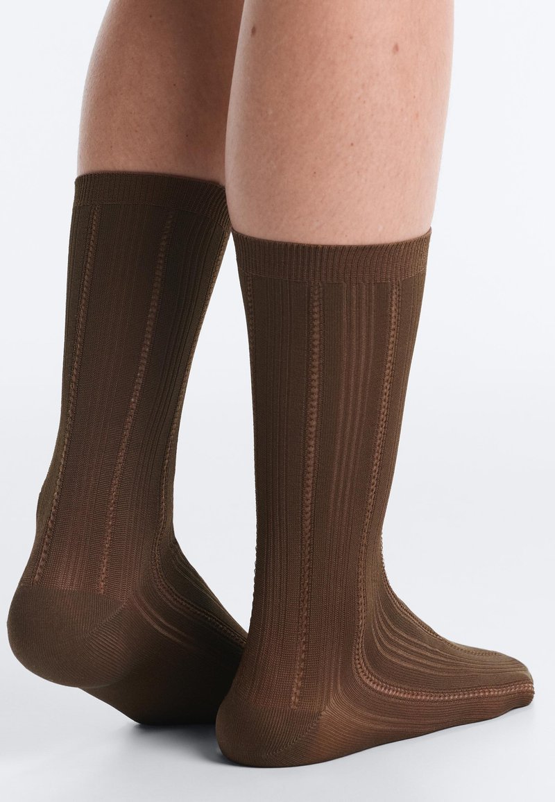 Jambes portant des chaussettes marron à motifs mi-mollet avec des rayures verticales côtelées et texturées, debout sur un fond clair.