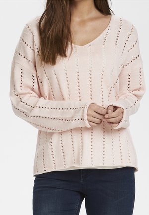 Kobieta w jasnoróżowym swetrze na drutach z pionowymi paskami z oczkami i ciemnoniebieskich dżinsach, z rękami złączonymi przed sobą.