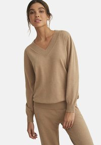 Pull en maille beige avec un col en V, des poignets et un ourlet côtelés. Le tissu semble doux et texturé, avec une coupe ample et un design épuré.