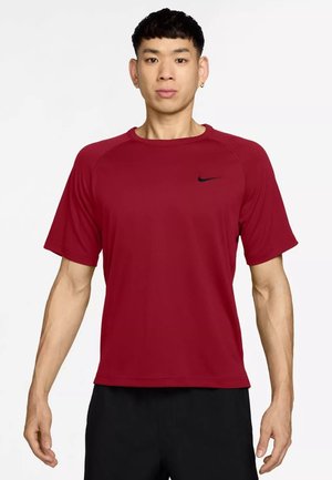 Uomo in piedi rivolto in avanti indossa una maglietta Nike rossa semplice a maniche corte e pantaloni neri su sfondo bianco.