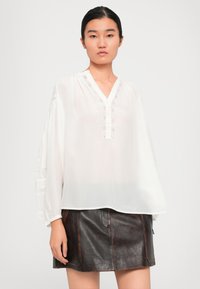 Jeune femme portant une blouse blanche ample avec des détails en dentelle et une mini-jupe en cuir marron foncé devant un fond clair.