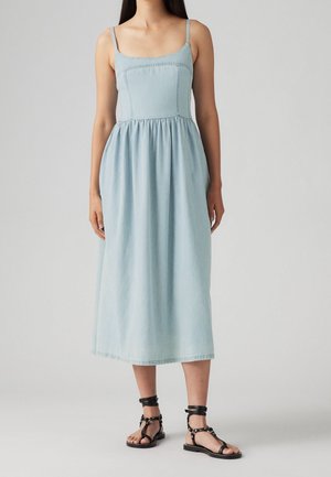 Kvinde iført en lyseblå, ærmeløs midi-kjole med indsnævret talje og sorte flade sandaler med ankelrem, stående foran en ensfarvet baggrund.