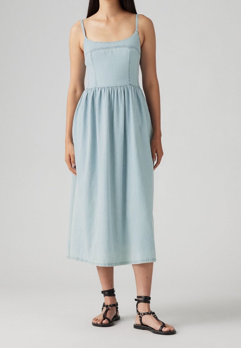 Femme portant une robe midi sans manches bleu clair à taille plissée et des sandales plates noires à bride cheville, se tenant contre un fond uni.
