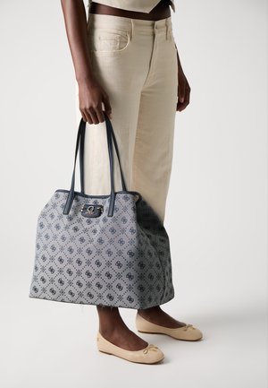 Sac tote gris à motifs avec des accents bleu foncé, présentant un détail de logo et des poignées doubles, associé à un pantalon large de couleur claire.
