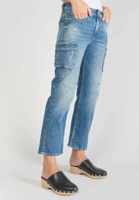 Mujer vestida con jeans cargo de mezclilla desgastados de color azul claro y zuecos de cuero negros con tacones de madera, de pie con las manos en los bolsillos.