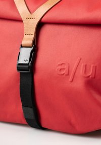 Borsa di stoffa rossa con una tracolla in pelle beige e una fibbia in plastica nera, con dettagli in rilievo del logo sul davanti per un tocco di texture.