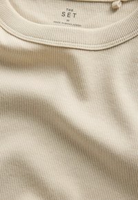 Beige ribbed cotton t-shirt collar with printed label stating "THE SET M Made in Bangladesh" on the inside back neckline.