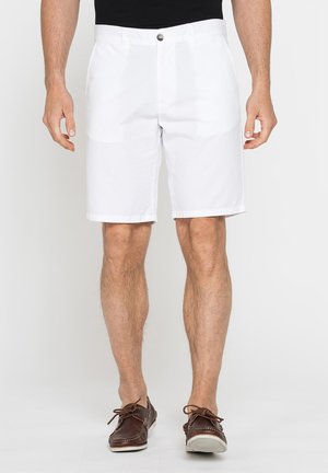 Uomo che indossa pantaloncini bianchi fino al ginocchio e scarpe da barca in pelle marrone, in piedi davanti a uno sfondo semplice.