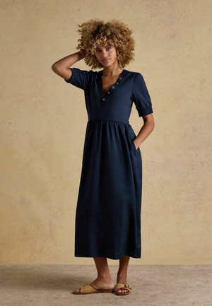Kobieta o kręconych włosach stoi boso, ubrana w granatową sukienkę midi z guzikami i krótkimi rękawami, jedną ręką trzyma się za głowę, drugą ma w kieszeni.