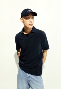 Marineblauwe polo met korte mouwen en v-hals, gemaakt van zacht materiaal, gecombineerd met lichtblauwe jeans en een bijpassende marineblauwe pet met tekst.