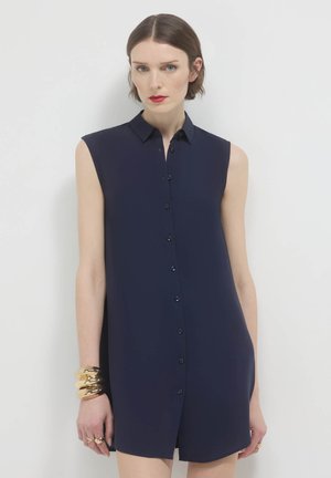 Abito camicia senza maniche di colore blu navy realizzato in tessuto liscio, con una chiusura davanti a bottoni e un colletto classico. Bracciali d'oro al polso.